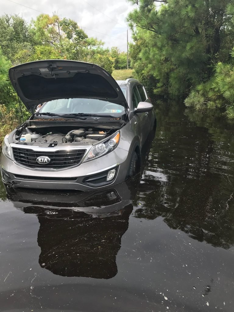 Gray Kia Sportage flooded with hood open in a dark, murky body of water surrounded by trees.
