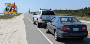 Silver SUV towing a blue sedan on a coastal road, North Beach Recovery logo visible.